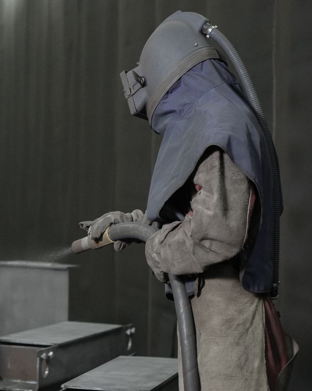 Arbeiter in Schutzkleidung bei der Bearbeitung von Metall mit einem Sandstrahler.