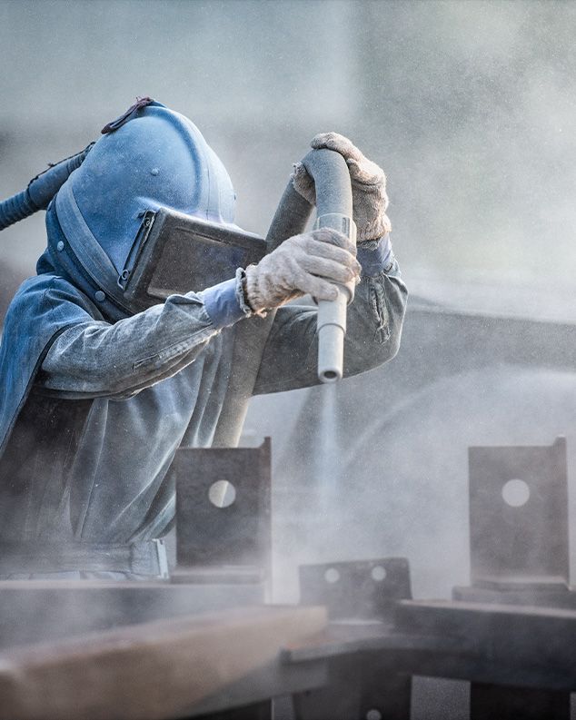 Arbeiter in Schutzkleidung bei der Bearbeitung von Metall mit einem Sandstrahler.